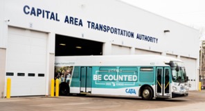 A picture of a bus outside a transportation warehouse. The bus is wrapped with "Be Counted" against a blue background.