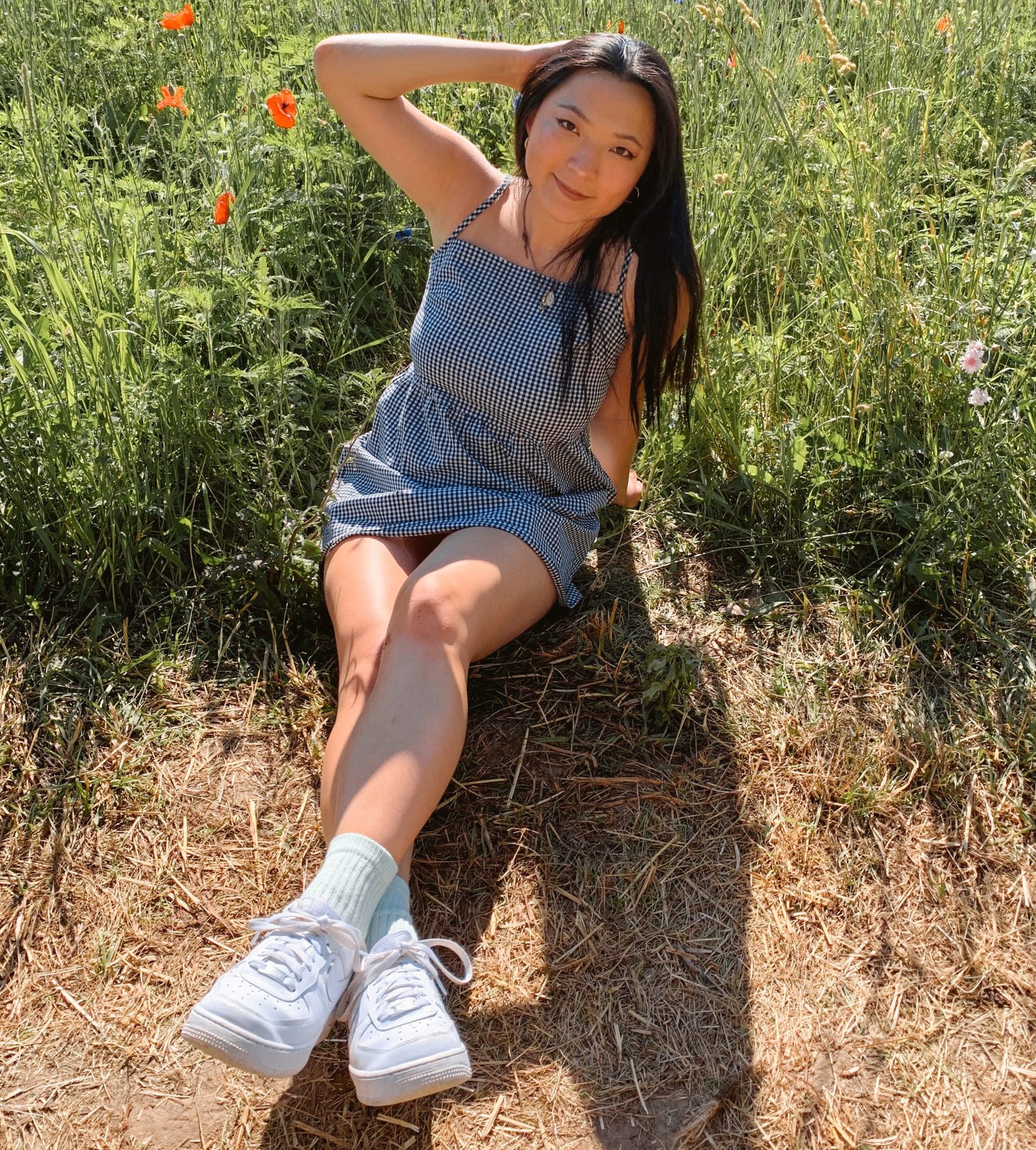 young woman with long black hair sitting in a field in overalls