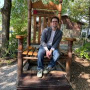 A exterior photograph of a man smiling, sitting on an oversized wooden chair.
