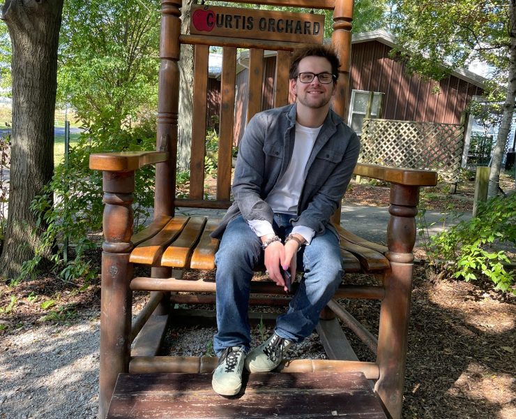 A exterior photograph of a man smiling, sitting on an oversized wooden chair.