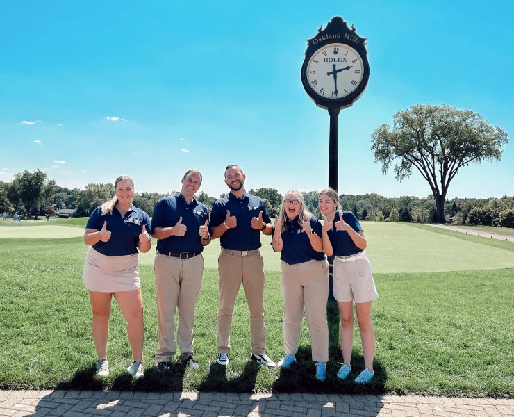 An exterior photograph of five students smiling near a large clock.