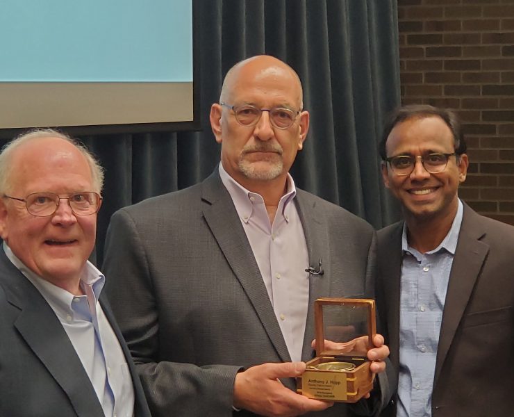 Three people pose while smiling. One person holds an award.