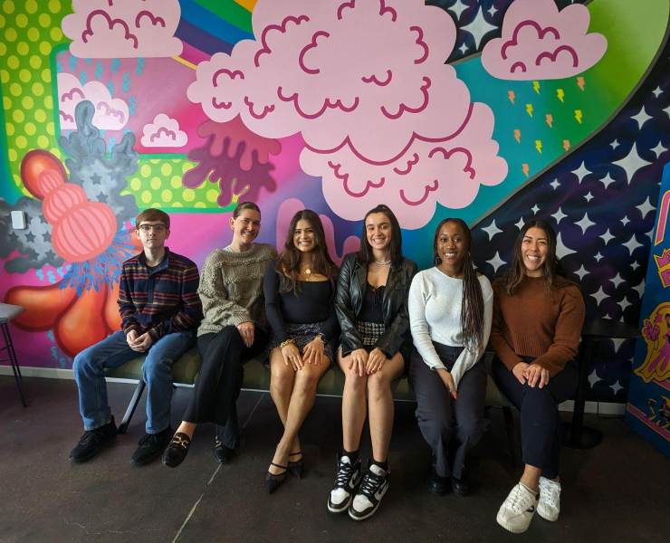 Six people smile while sitting for a photo in front of a multi-colored mural.