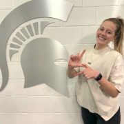 An MSU student poses in front of a wall with a MSU Spartan Helmet logo