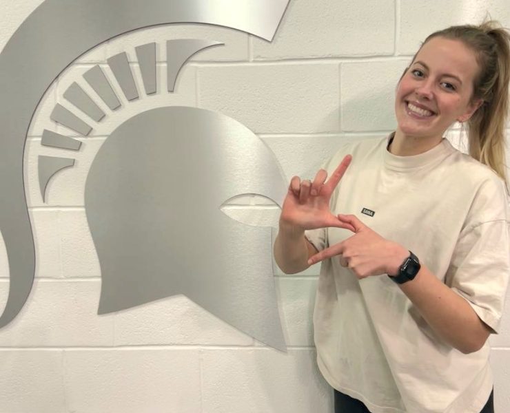 An MSU student poses in front of a wall with a MSU Spartan Helmet logo