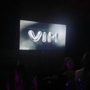 A photograph of a presentation with lights down. The screen shows the letters VIM in shiny silver lettering