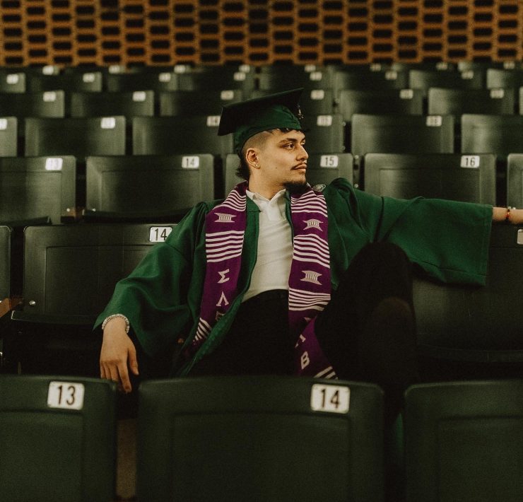A person sitting in graduation attire in a seats in a classroom