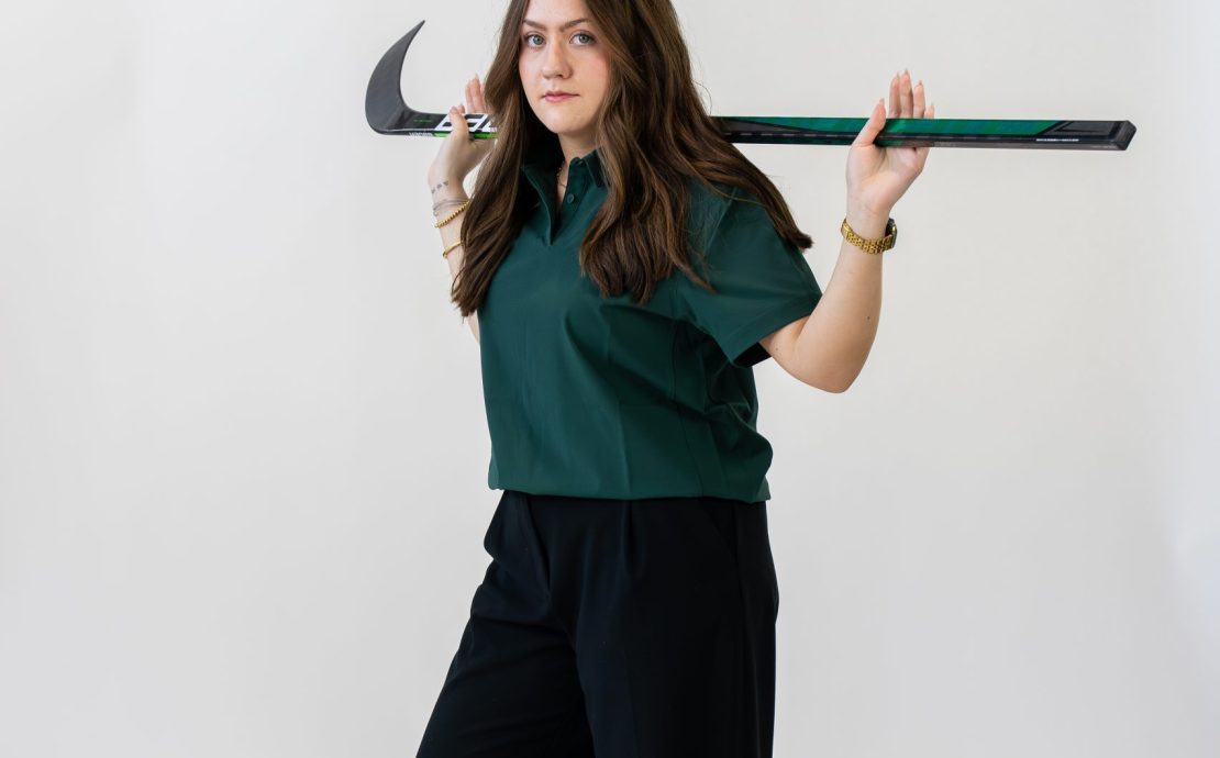 A person poses on a white background with a hockey stick