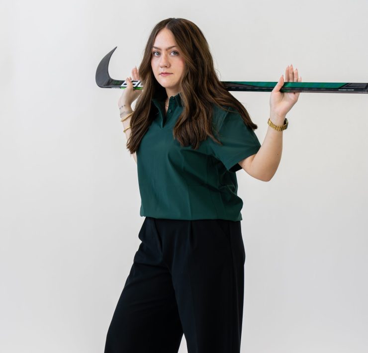A person poses on a white background with a hockey stick
