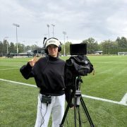 A woman gives a thumbs up next to a broadcast camera on a football field